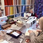 Three women focused on their sewing machines during a crafting workshop inside The Patchwork Basket unit at The Hawthorns Business Centre, surrounded by shelves full of colorful fabric bolts.