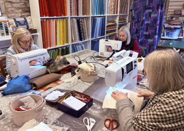 Three women focused on their sewing machines during a crafting workshop inside The Patchwork Basket unit at The Hawthorns Business Centre, surrounded by shelves full of colorful fabric bolts.