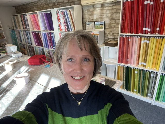 Portrait of Mags Smith, owner of The Patchwork Basket, smiling inside her commercial workspace at The Hawthorns Business Centre, with rainbow-organised fabric rolls displayed on white shelving