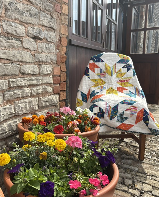 A vibrant patterned quilt draped over a chair on a cobbled courtyard outside The Hawthorns Business Centre, flanked by stone walls and pots of blooming marigolds and geraniums.
